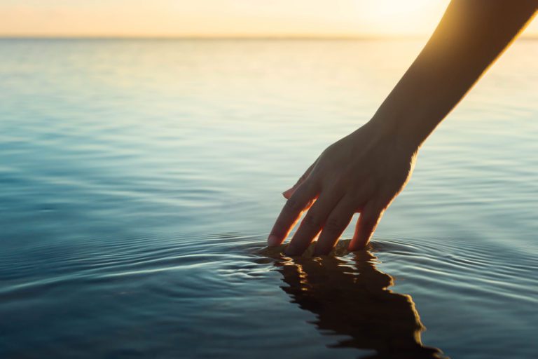 female hand touching the ocean water in front of a beautiful sunset